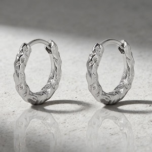 May include: A pair of silver-colored hoop earrings. Each earring has a textured, organic design. The earrings are displayed on a light-colored, reflective surface, with shadows adding depth. The earrings are a jewelry item.