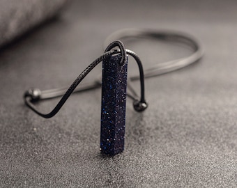 Op de afbeelding: Een zwarte koordketting met een donkerblauwe, rechthoekige hanger. De hanger heeft een glinsterende, gespikkelde textuur.