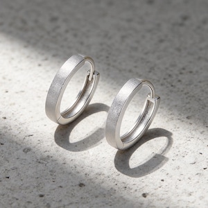 May include: A pair of silver hoop earrings with a matte, textured finish. The earrings feature a hinged closure mechanism and a circular design. The metal surface has a subtle, granular appearance that catches the light, creating a soft contrast.