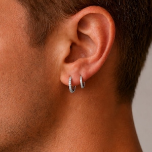 May include: Two small, silver hoop earrings, each encrusted with small, clear gemstones. The earrings are worn on the ear of a person with short, dark hair. The earrings are of different sizes, with the larger one closer to the earlobe.