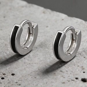 May include: Two silver hoop earrings with a black stripe detail. The earrings are shown on a textured gray surface. The earrings have a simple, modern design.