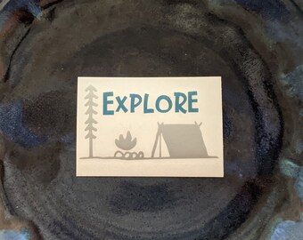 Könnte beinhalten: Eine beige Karte mit einer blauen Silhouette eines Baumes, eines Lagerfeuers und eines Zeltes. Der Text "EXPLORE" ist in blauen Buchstaben geschrieben.