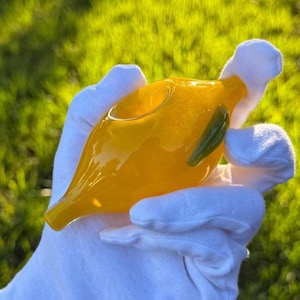 May include: A hand-blown glass pipe in a vibrant yellow hue, featuring a rounded bowl and a slender, tapered stem. A small, green glass accent is visible on the side. The pipe is held in a gloved hand against a backdrop of green grass.