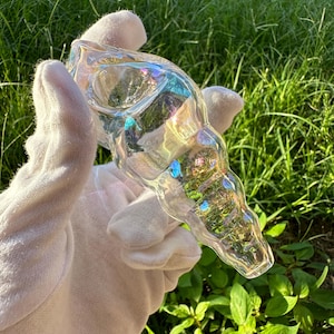 May include: A clear glass seashell pipe with iridescent rainbow colors. The pipe is shaped like a seashell and has a small bowl at the top.