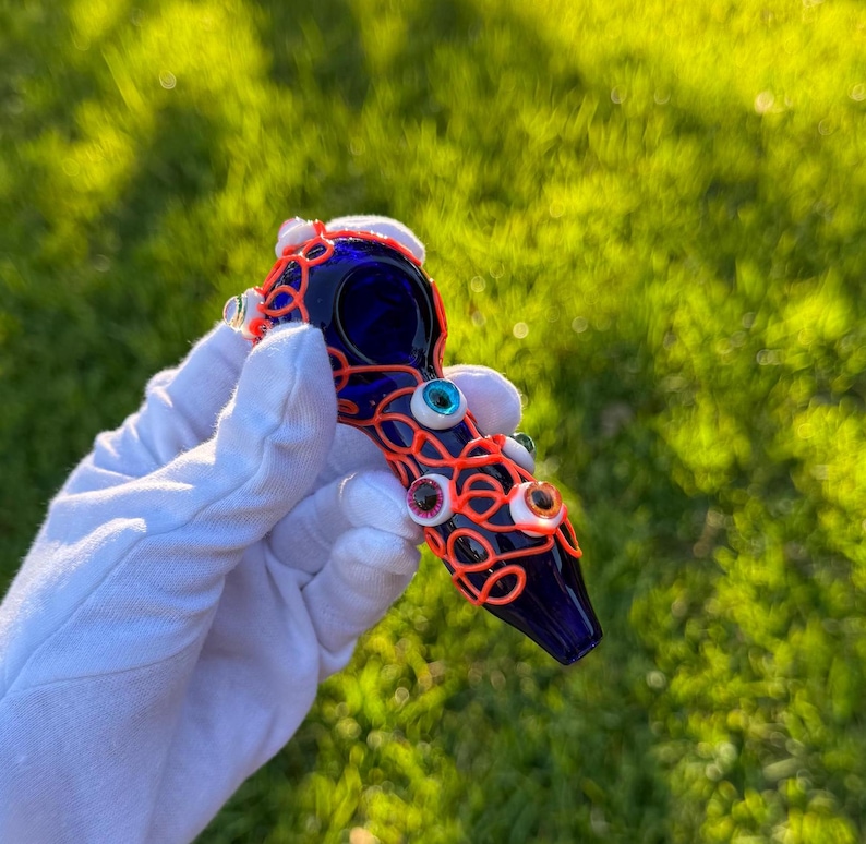May include: A vibrant blue glass smoking pipe adorned with an intricate orange wire design.  The wire is interwoven with several small, colorful plastic eyeballs, adding a whimsical, quirky touch. The pipe's bowl is deep blue, contrasting with the bright orange wire. This unique piece is a collectible item, perfect for enthusiasts of artistic smoking accessories.