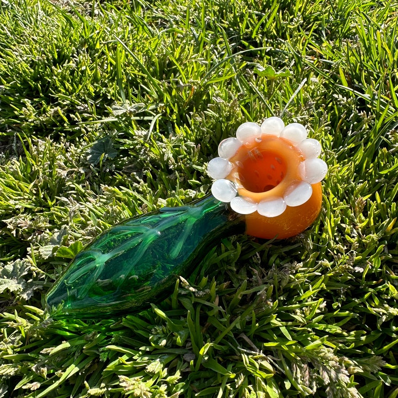 May include: A green glass pipe with a white and orange flower-shaped bowl. The pipe is lying on a bed of green grass.