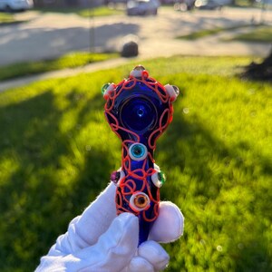 May include: A blue glass pipe with a red and white swirl design and colorful eyes. The pipe is decorated with a variety of colorful glass eyes.