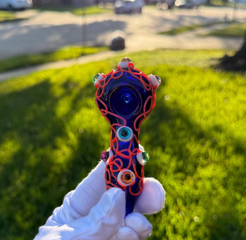 May include: A blue glass pipe with a red and white swirl design and colorful eyes. The pipe is decorated with a variety of colorful glass eyes.