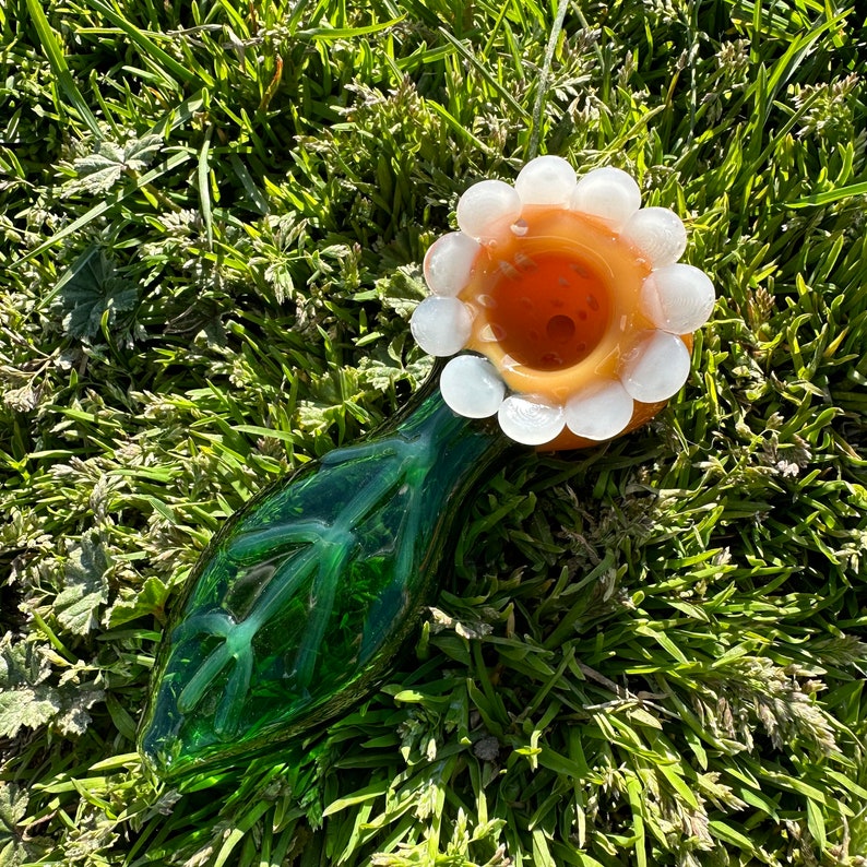 May include: A green glass pipe with a white and orange flower-shaped bowl. The pipe is designed to resemble a flower with a stem and leaves.