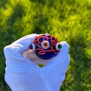 May include: A blue glass orb with a red wire design and three eyes. The eyes are made of glass and have different colors, including orange, white, and green.