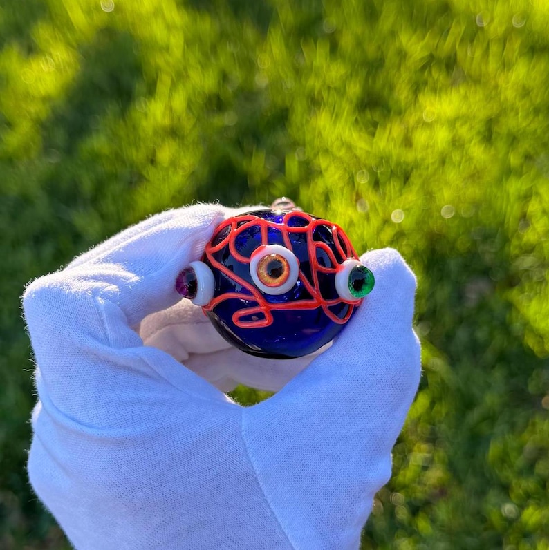 May include: A blue glass orb with a red wire design and three eyes. The eyes are made of glass and have different colors, including orange, white, and green.