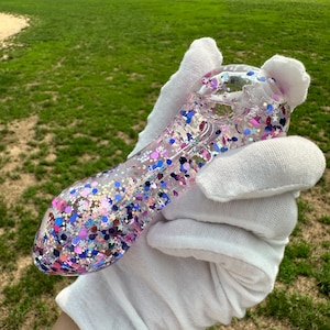 May include: A clear glass pipe filled with colorful glitter in shades of pink, blue, purple, and silver. The pipe is held in a white gloved hand against a green grassy background.
