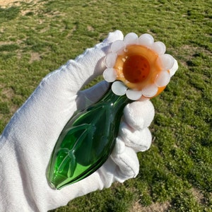 May include: A green glass pipe with a flower-shaped bowl. The bowl is orange with white accents around the rim.