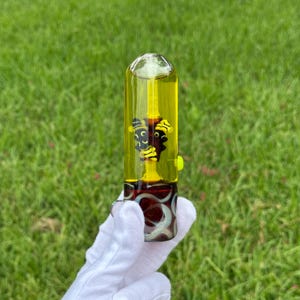 May include: A clear glass smoking pipe with a yellow liquid inside. The pipe has a decorative design in the center and a patterned base. The pipe is held by a gloved hand against a green grass background.