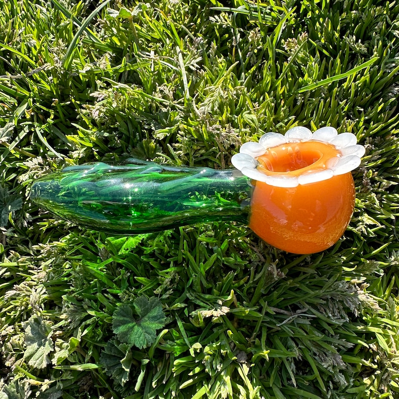 May include: A green glass pipe with a swirl design and an orange bowl with a white daisy-shaped rim.