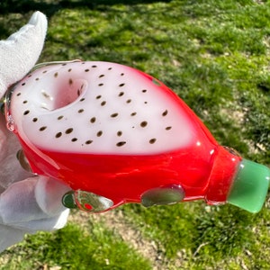 May include: A red and white glass pipe shaped like a dragon fruit. The pipe has a green mouthpiece and black dots on the white part.