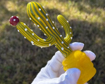 May include: A glass cactus sculpture with green and yellow stripes and white dots. The cactus has a red flower on top and a yellow base.