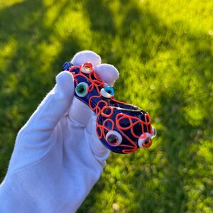 May include: A blue glass pipe with a red and orange swirl design. The pipe is decorated with colorful glass eyes.