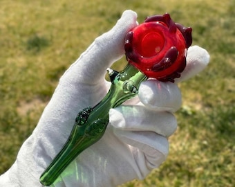 May include: A glass pipe shaped like a rose, featuring a vibrant red bowl at the top and a translucent green stem with sculpted leaf details. The piece is crafted from smooth, polished glass, showcasing a floral design with a deep red flower head.