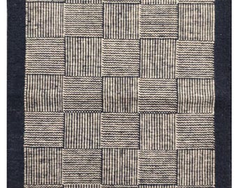 May include: A rectangular rug with a woven checkerboard pattern. The design features alternating squares of vertical stripes and solid blocks in shades of cream and navy blue. The rug has a navy blue border and fringe.