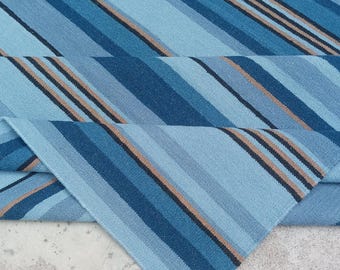 Op de afbeelding: Een gevouwen blauw en bruin gestreept tapijt. Het tapijt heeft verschillende tinten blauw, met bruine en zwarte strepen. Het tapijt is gemaakt van een geweven materiaal en is op zichzelf gevouwen, waardoor de onderkant zichtbaar is.