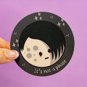 Peut inclure: Un autocollant noir et blanc avec une lune de dessin animé avec une coiffure emo noire. La lune a une expression triste et des joues rouges. L'autocollant porte le texte "Ce n'est pas une phase".