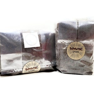 May include: Two clear plastic bags filled with dark brown mushroom spores. The bags have a brown label with the text "Seismic Spore" printed on them.