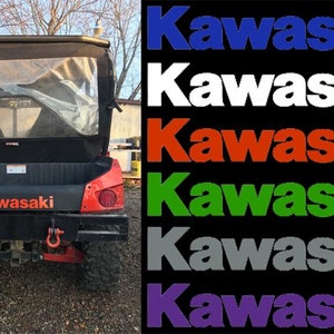 May include: A red Kawasaki utility vehicle with a black canvas top and rear window. The vehicle has a black bed with the word "Kawasaki" printed in white. The vehicle is parked on a gravel surface. The image also includes six different color variations of the word "Kawasaki" in blue, white, red, green, gray, and purple.