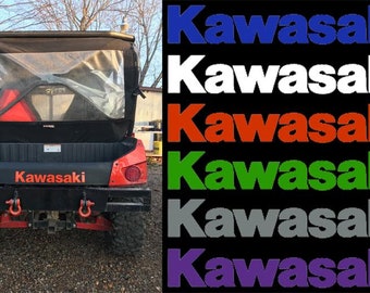 May include: A red Kawasaki utility vehicle with a black canvas top and rear window. The vehicle has a black bed with the word "Kawasaki" printed in white. The vehicle is parked on a gravel surface. The image also includes six different color variations of the word "Kawasaki" in blue, white, red, green, gray, and purple.