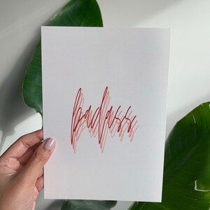 Puede incluir: Una tarjeta blanca con la palabra "badass" escrita en cursiva roja.