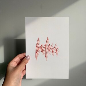 Puede incluir: Una tarjeta blanca con la palabra "badass" escrita en cursiva roja y rosa.