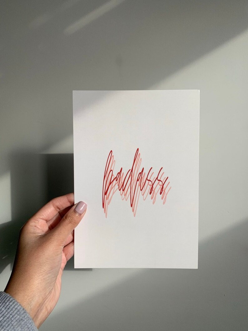 Puede incluir: Una tarjeta blanca con la palabra "badass" escrita en cursiva roja y rosa.