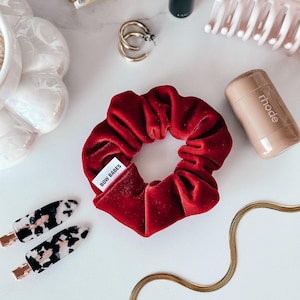 Può includere: Un elastico per capelli in velluto rosso con un'etichetta bianca che dice "BOW BABES".