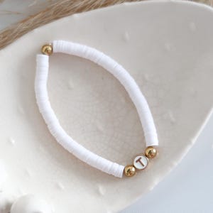 May include: White beaded bracelet with gold accents and a letter "T" charm. The bracelet is made of small, flat, white beads and gold-coloured round beads. The bracelet is displayed on a white, textured surface.