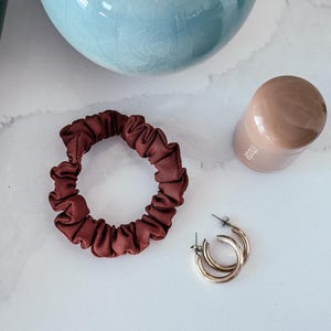 May include: A close-up of a brown scrunchie, gold hoop earrings, and a brown container on a white marble surface.