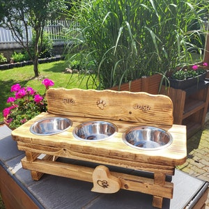 Può includere: Stazione per cibo per animali domestici in legno con tre ciotole in acciaio inox. La stazione ha un ritaglio a forma di cuore con un'impronta di zampa ed è incisa con i nomi "Ella" e "Joss".