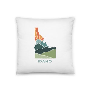 Könnte beinhalten: Ein weißes Kissen mit einer bunten Grafik des Bundesstaates Idaho. Die Grafik zeigt einen Sonnenaufgang über Bergen und das Wort "IDAHO" in blauer Schrift.