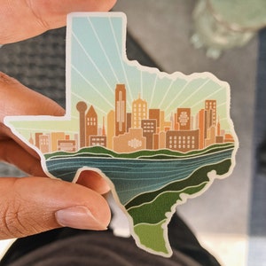 May include: A sticker shaped like the state of Texas with a skyline of buildings in the center. The buildings are brown and orange, and the sky is blue with white rays of light. The sticker is on a green and blue background.