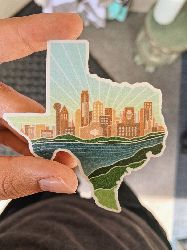 May include: A sticker shaped like the state of Texas with a skyline of buildings in the center. The buildings are brown and orange, and the sky is blue with white rays of light. The sticker is on a green and blue background.