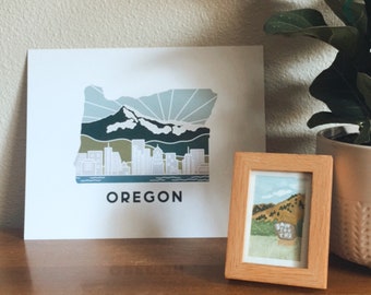 May include: A white print with a stylized illustration of the state of Oregon, featuring a mountain range and a cityscape. The print is on a wooden table with a small framed print of a landscape scene next to it.