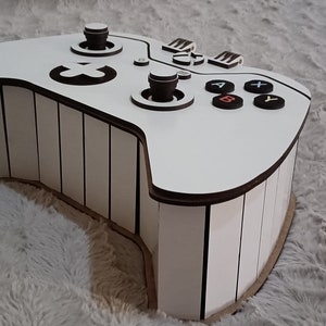 Puede incluir: Caja de madera blanca con forma de mando de Xbox con detalles negros. La caja tiene un diseño a rayas y presenta los botones estándar del mando de Xbox.