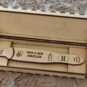 Puede incluir: Una caja de madera con una pequeña placa de madera en su interior. La placa tiene un chupete, un biberón y dos líneas, junto con el texto "VAIS A SER ABUELOS".