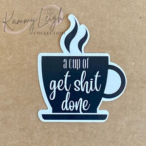 May include: A black and white sticker in the shape of a coffee cup. The text on the cup reads "a cup of get shit done" in a white, cursive font. The sticker has a steam design above the cup.