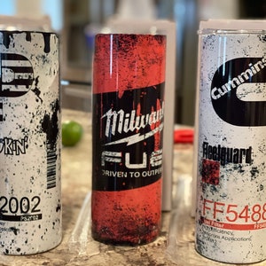 May include: Three stainless steel tumblers with different designs. The first tumbler has a white background with black splatter paint and the text "HOKIN PS2002". The second tumbler has a red background with black splatter paint and the text "Milwaukee FUEL DRIVEN TO OUTPER". The third tumbler has a white background with black splatter paint and the text "Cummins Reelguard FF5488 Fuel Filter Efficiency Microns Applications FF5488".