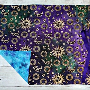 May include: A purple and green fabric square with a gold sun and moon pattern. The fabric has a tie on each side. The back of the fabric is a light blue color.
