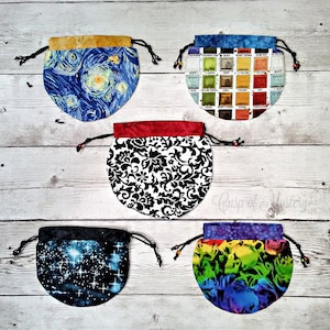 May include: Five drawstring bags with different patterns. The bags are made of fabric and have a round shape. The patterns include a starry night sky, a watercolor palette, a black and white damask pattern, a galaxy with stars, and a rainbow skull pattern.