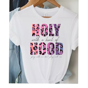 May include: White t-shirt with a pink, purple, and blue leopard print design that says "HOLY with a hint of HOOD pray with me don't play with me".