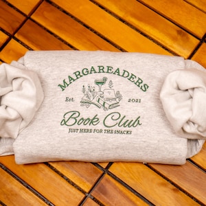 May include: A light heather gray sweatshirt featuring dark green embroidery that reads MARGAREADERS BOOK CLUB, Est. 2021, and JUST HERE FOR THE SNACKS. The design includes illustrations of a cocktail glass, open books, and floral sprigs.
