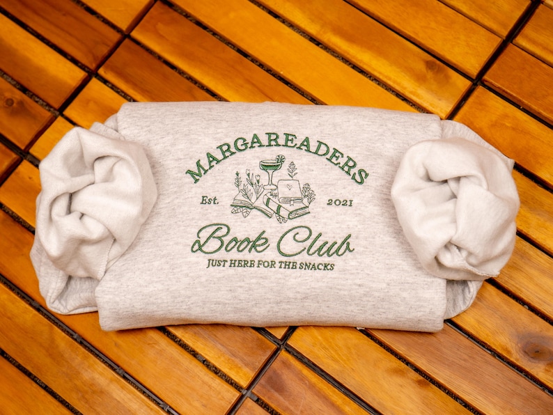 May include: A light heather gray sweatshirt featuring dark green embroidery that reads MARGAREADERS BOOK CLUB, Est. 2021, and JUST HERE FOR THE SNACKS. The design includes illustrations of a cocktail glass, open books, and floral sprigs.