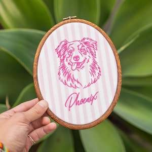 May include: An oval wooden embroidery hoop containing white fabric with vertical light pink stripes. The fabric features a magenta embroidered portrait of a dog with its tongue out, positioned above the cursive magenta text reading Phoenix.